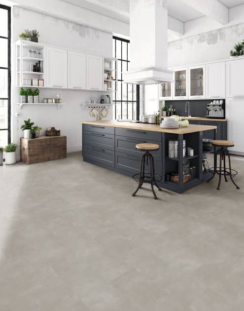 Industrial-inspired white and blue kitchen with large windows, hanging wall cabinets, rustic chest, potted houseplants, and two stools, and light gray stone concrete-look LVT