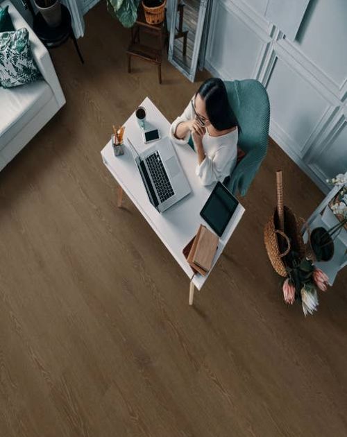 Woman works on laptop on desk in home office, beside sofa, basket of flowers, and shelf, with wainscoting up walls and medium brown woodgrain flooring