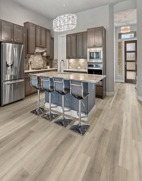 Kitchen with neutral brown cabinets, pale blue accent island with chairs, with wide neutral brownish gray woodgrain flooring, and entry door visible in background