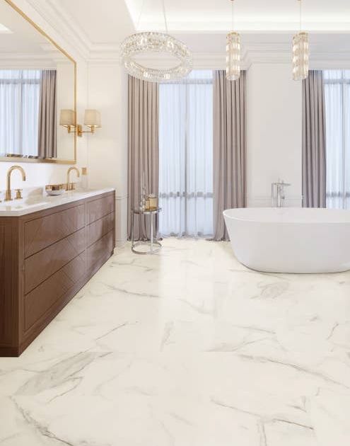 Luxury bathroom with tall windows, multiple chandeliers, tub, wooden vanity, gold mirror and fixtures, and white-and-gold marble stone LVT floor