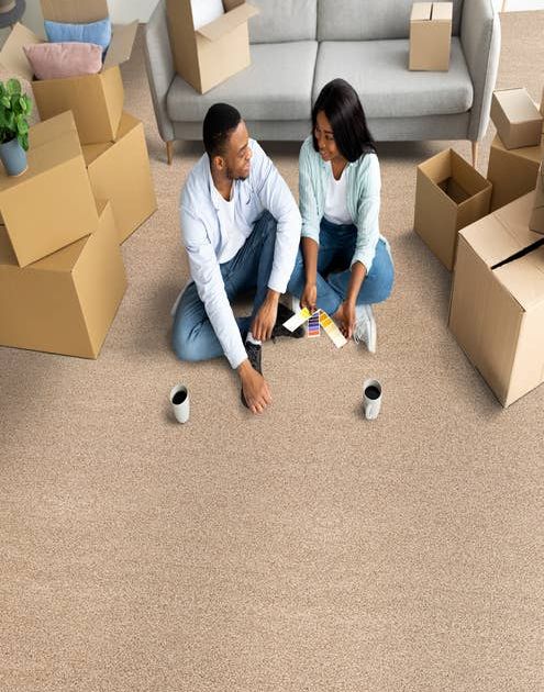 Young couple sitting on solid texture tan carpet with coffee mugs and moving boxes