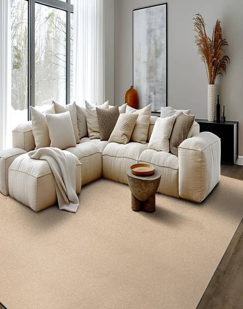 Cozy living room scene with a modern plush cream couch topped with an array of pillows and a throw blanket while snow falls outside