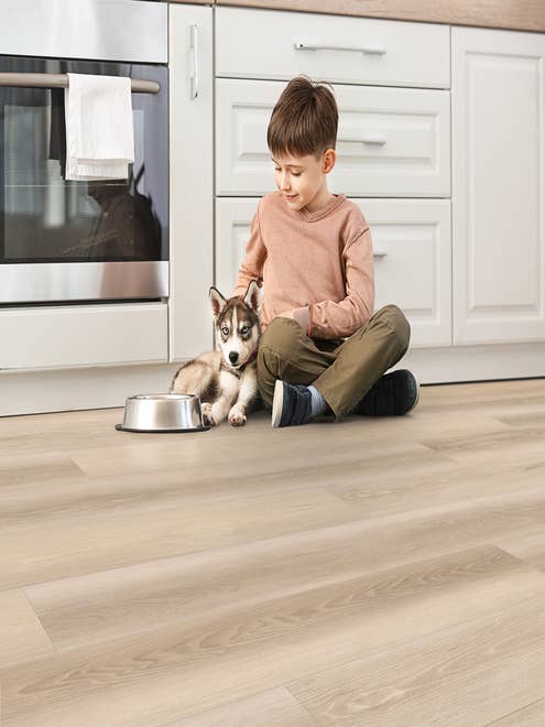 Young boy and husky puppy sit on kitchen floor before white cabinets and light woodgrain vinyl flooring