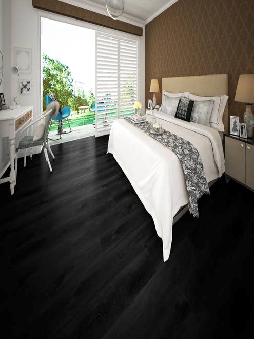 Room with brown patterned wallpaper, black LVP floors, inviting bed, desk, and sliding door leading to deck space with blue chairs