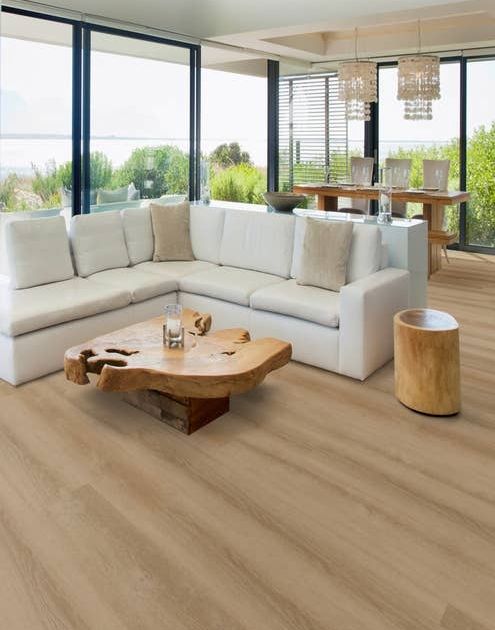 Beachy living room surrounded by floor-to-ceiling windows, with cozy white sofa, driftwood coffee table, and taupe woodgrain floor, with dining area and twin chandeliers