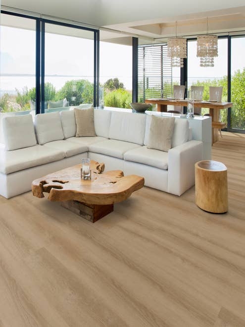Beachy living room surrounded by floor-to-ceiling windows, with cozy white sofa, driftwood coffee table, and taupe woodgrain floor, with dining area and twin chandeliers