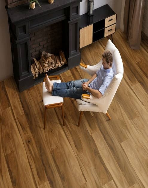 Man in modern, organic armchair sits with feet up on ottoman across from black fireplace, in room with dramatic and rich hickory-look floor