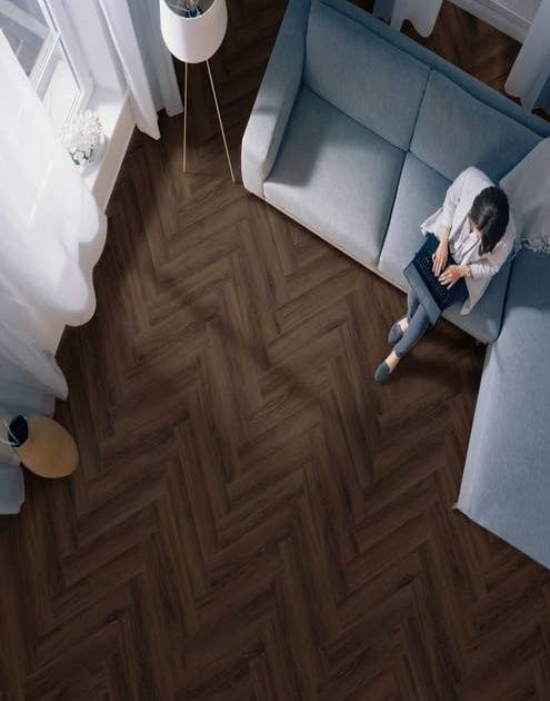 Woman on laptop sits on blue couch in dark and moody living room with dramatic, rich brown herringbone floors