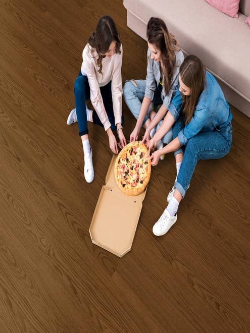 Three teenage girls sit on dark, rich brown LVP floor and share a box of pizza