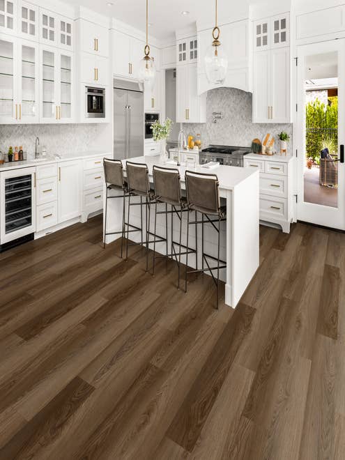 White kitchen cabinetry with gold accents, gold and glass chandeliers, and white island with barstools over Old World Almond Bark LVP flooring