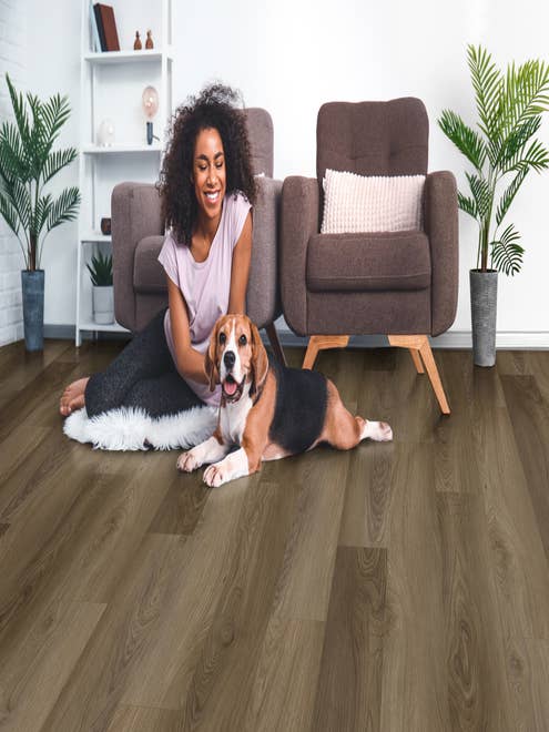 Woman sits in front of two mid-century armchairs and pets dog, over tonal brown Hazelnut Harvest LVP