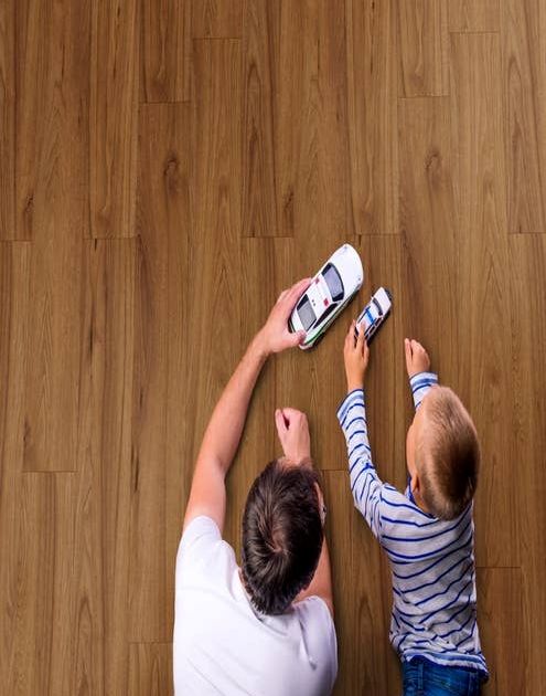 Man and boy lay on rich, reddish brown laminate floor and play with toy cars