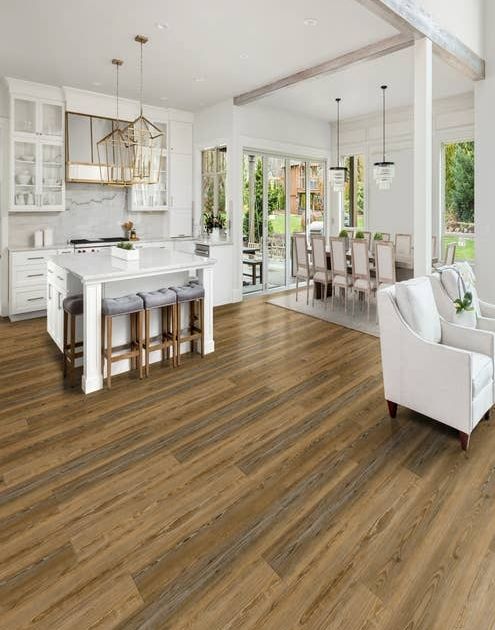 Open-concept kitchen, dining, and seating areas, with white walls, gold accents, and brown naturally-aged luxury vinyl plank floor