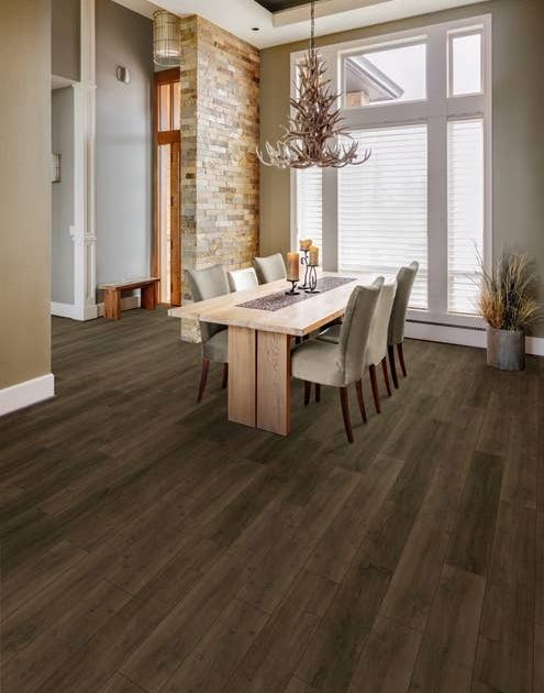 Dining area with table and chairs beneath antler chandelier, in room with exposed brick wall, large window, and entryway in background, with medium to dark brown LVP flooring