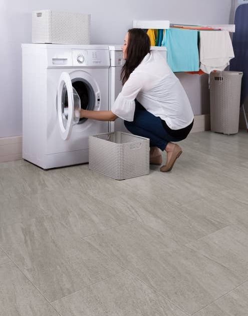 Woman crouches before washing machine and does laundry, next to hanging wrack and hampers, in laundry room with gray walls and gray stone-look LVT floor