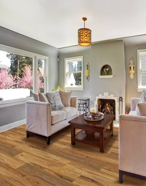 Cozy, calm living room with large windows, golden brown floors, two couches facing a wooden coffee table before a fireplace, with sconces and a patterned chandelier