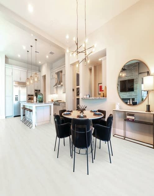 Open concept kitchen and dining area, with white cabinetry, island with barstools, and pendant lighting, along with table and chairs under large chandelier, before round mirror, in room with tall ceilings and white woodgrain LVP floors
