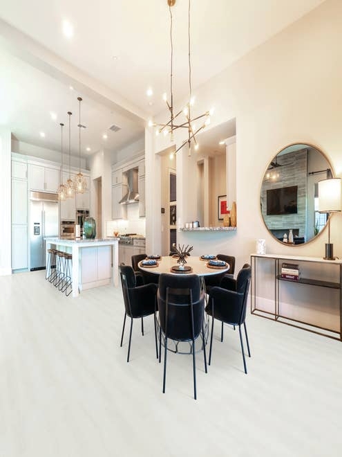 Open concept kitchen and dining area, with white cabinetry, island with barstools, and pendant lighting, along with table and chairs under large chandelier, before round mirror, in room with tall ceilings and white woodgrain LVP floors