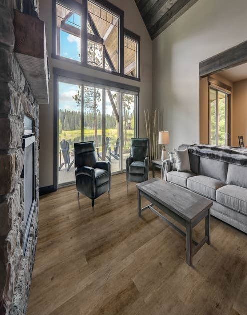 Cabin with large fieldstone fireplace, two armchairs, plush gray sofa, and coffee table, with large windows overlooking patio with Adirondack chairs