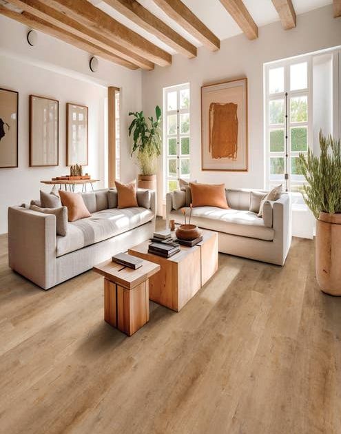 Bright living room with large windows, artwork, two plush sofas facing a coffee table, and houseplants, with exposed wood beams and a tan woodgrain floor
