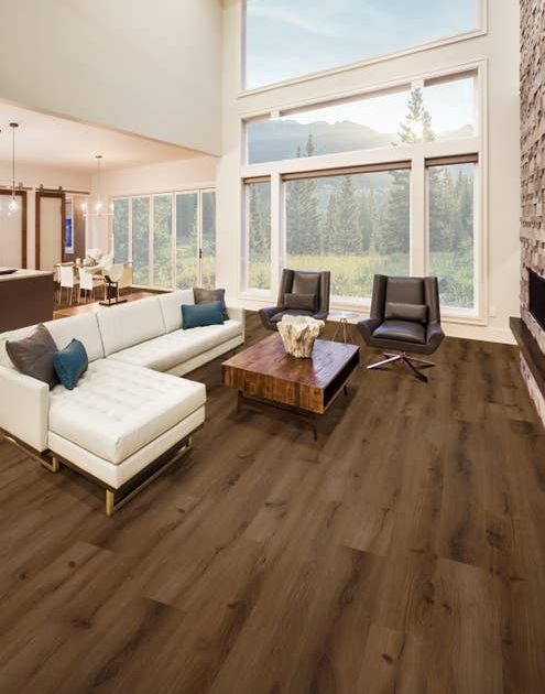 Contemporary open concept living room with medium brown wood-look floors with white sectional sofa, two armchairs, coffee table, large stone fireplace, and several large windows showing wilderness beyond