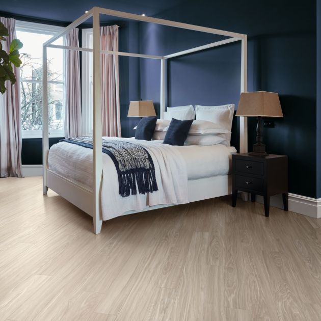 Calm bedroom with dark blue walls, gray woodgrain floors, cozy bed, night stands, large plant, and window