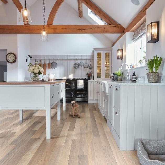 Kitchen with brown woodgrain floor, and white cabinetry, island, with exposed arched wooden beams, and various cooking equipment hanging on wall, with dog sitting before stove