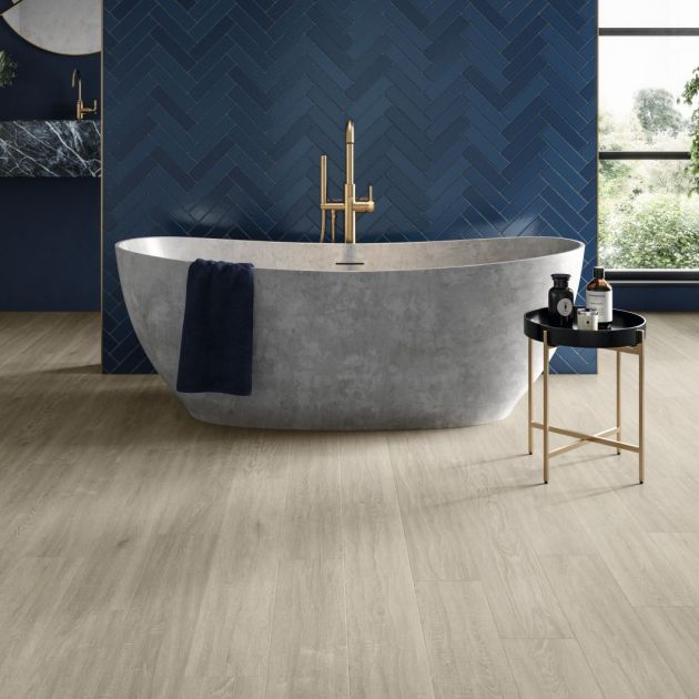 Moody bathroom with taupe woodgrain floor, with large gray soaker tub and table before dark blue herringbone tile wall, with window, potted plant, and sink in background