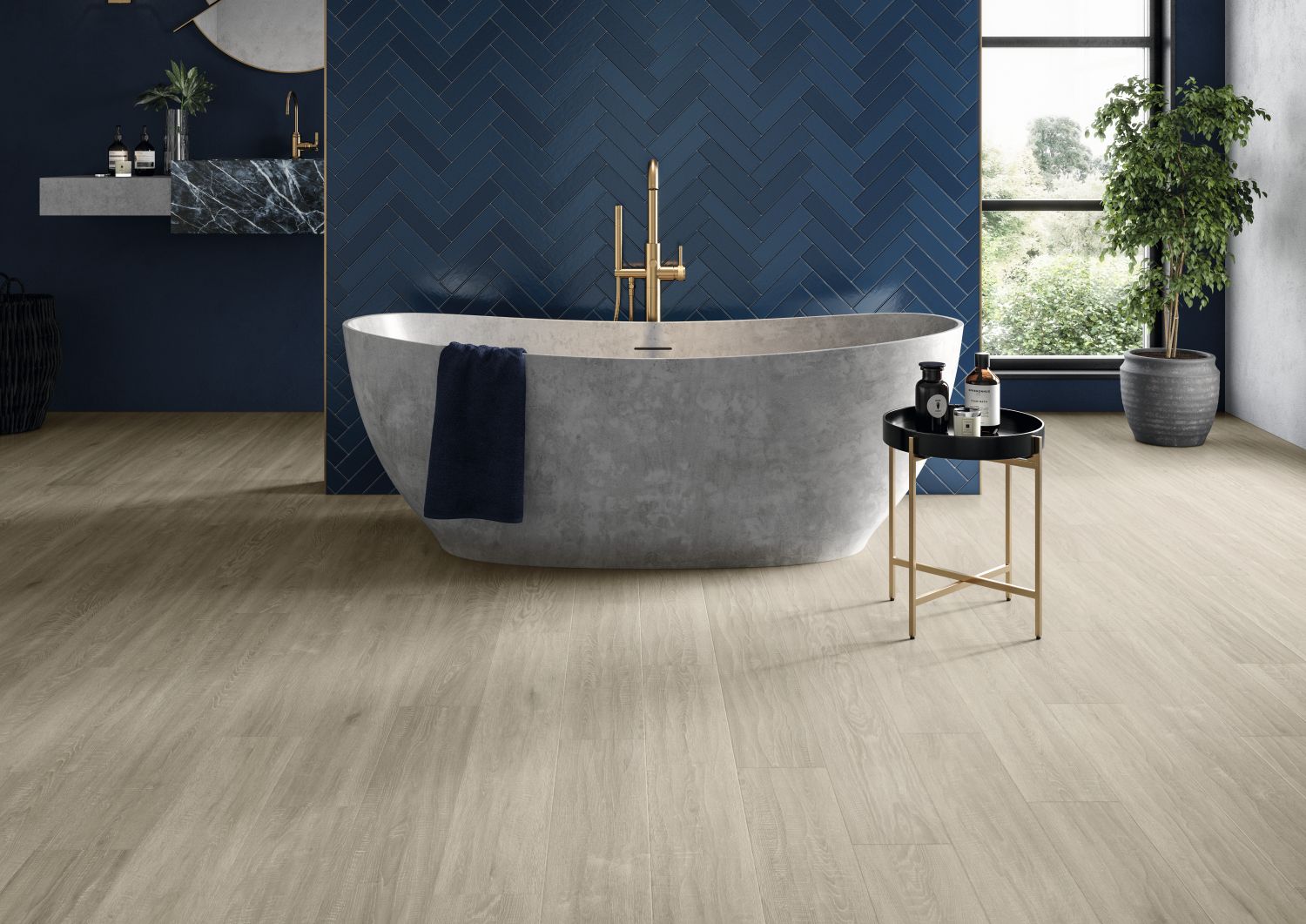 Moody bathroom with taupe woodgrain floor, with large gray soaker tub and table before dark blue herringbone tile wall, with window, potted plant, and sink in background