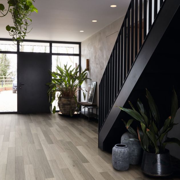 Dramatic entry hallway featuring black staircase, black door surrounded by windows, limestone washed walls, gray woodgrain floor, and several large plants and ceramic vessels, with recessed lighting and glimpse of upper floor