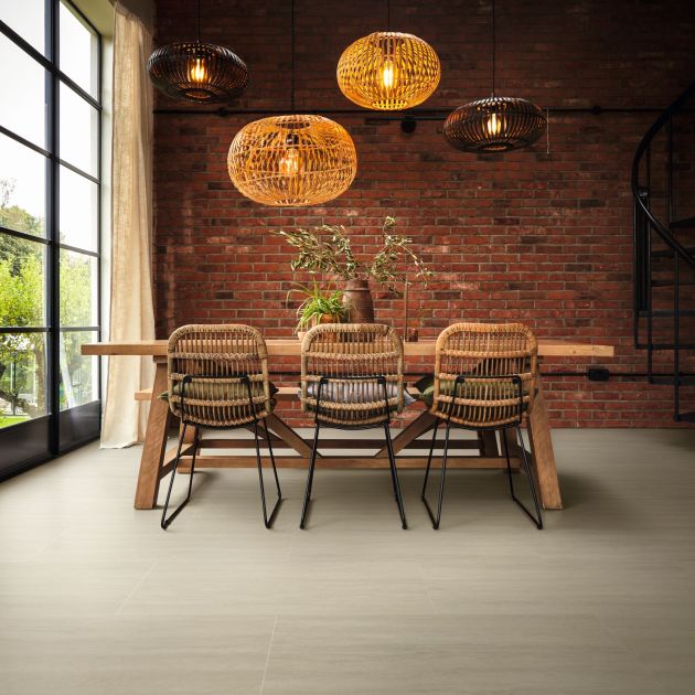 Modern dining table and wicker chairs under multiple woven light fixtures, with exposed brick wall, large black trimmed windows flanked by curtains, iron spiral staircase, and Travertine stone LVT floor