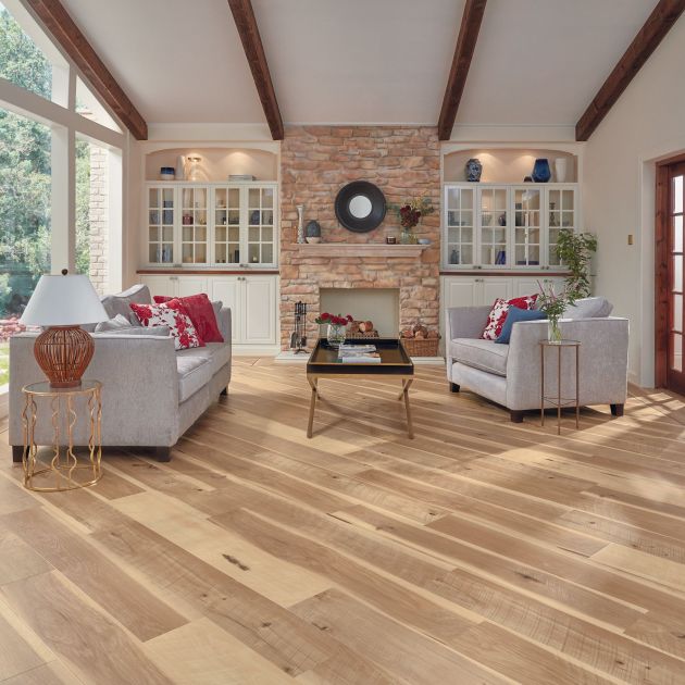 Bright living space with exposed wooden beams, brick fireplace, white built-in cabinets, large window, and contemporary sofa facing coffee table and armchair, with gold metal end tables, over distressed wide-plank hickory woodgrain floor