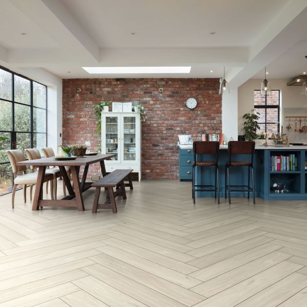 Open-concept kitchen with blue cabinetry, island, and open shelving, and pendant lighting, leading to dining area with table, bench, and chairs before exposed brick wall with China cabinet and large windows, and cream herringbone woodgrain floor