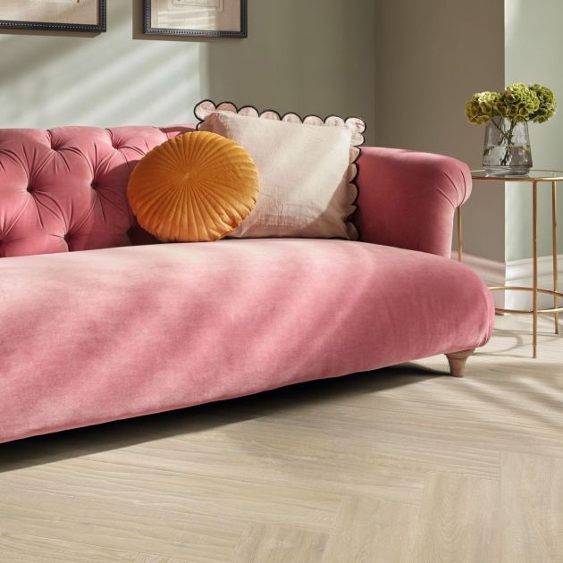 Pink velvet tufted couch and cushions beside gold metal end table, under picture frames, with herringbone woodgrain floor