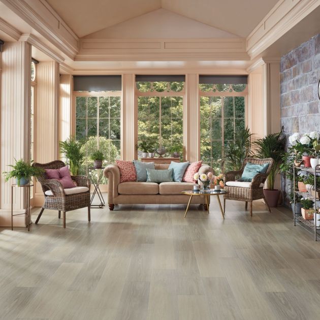 Sunroom with grand windows between columns, exposed brick wall, sofa and wicker chairs, and shelf filled with potted plants, with various other potted plants, with clean gray woodgrain floor
