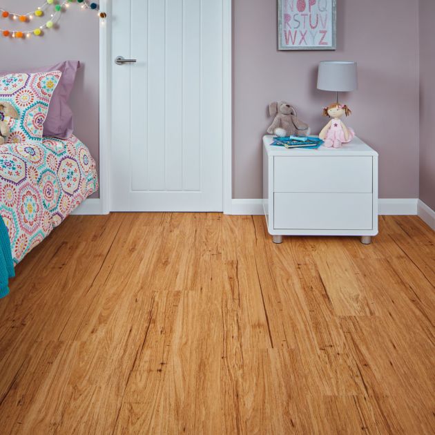 Girl's bedroom with lilac walls, white door, golden woodgrain floor, vibrant floral bedspread, and white nightstand with stuffed animals and lamp under alphabet artwork