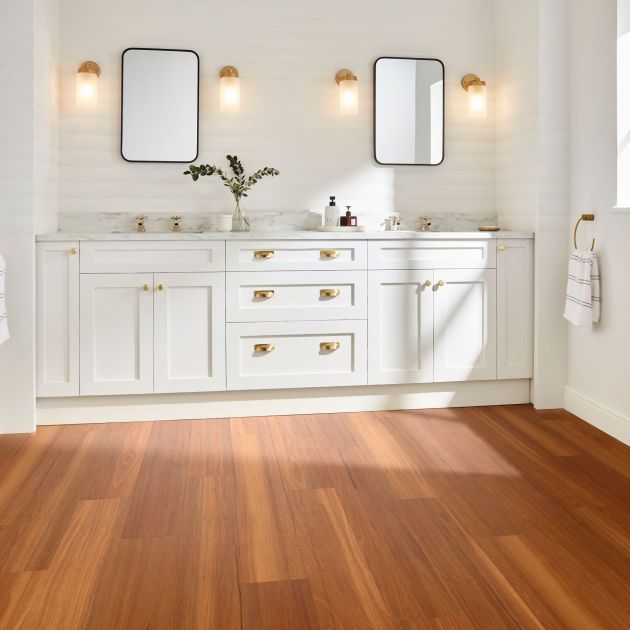 White bathroom dual sink vanity with gold fixtures, sconces, and window with sunlight shining on orange and brown woodgrain floor