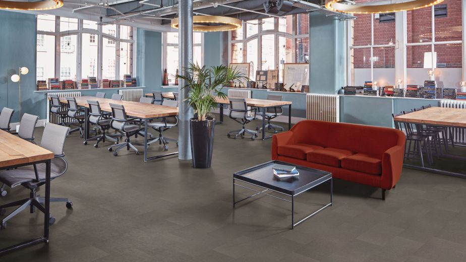Open-concept commercial office space with gray tile floors, oversized light fixtures, meeting desks, red velvet couch, and coffee table, with view of brick building outside