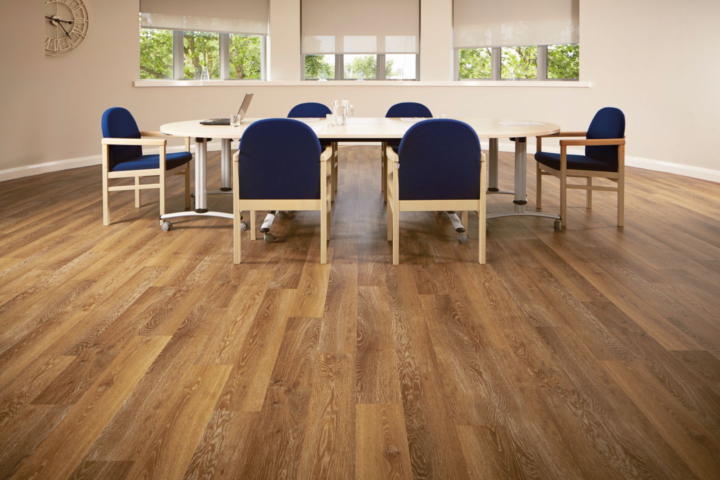 Office space with taupe walls and copper-toned wood floor, with large clock and bank of windows overlooking meeting table and chairs