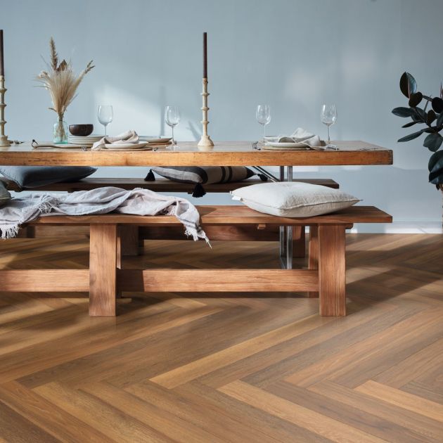 Wooden dining table and bench with wine glasses, candlestick, and pillows over herringbone pattern golden brown LVP floor, with potted plant in corner
