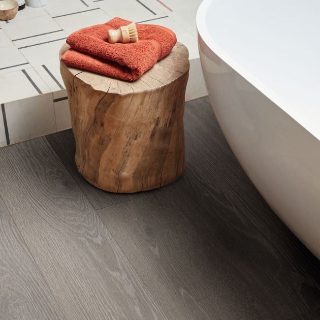 Bathtub and stump side table topped with towel and shave brush, with white and gray geometric shower floor in background, with medium gray woodgrain floor