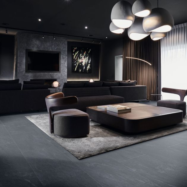 Dark, bold modern living area with large chandelier, TV, and artwork, with couch and chairs around coffee table on area rug and dark tile floor
