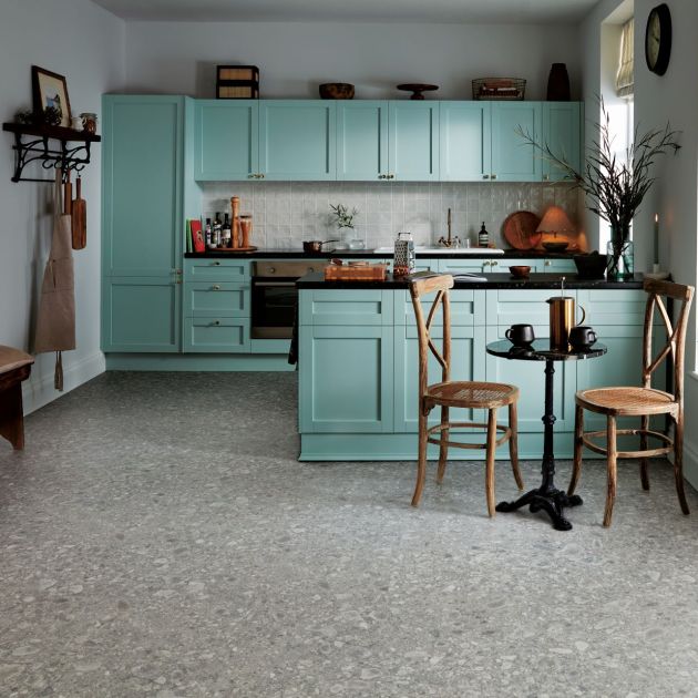 Icy blue kitchen cabinetry and island facing small eating area, with floating shelf and various paraphernalia around, with gray terrazzo-like stone tile floor