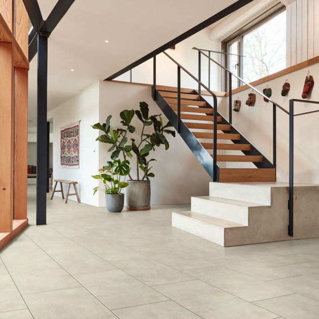 Modern living space and hall with stone steps leading to open wood and metal staircase, various hanging animal masks below large window, house plants, hanging tapestry and bench, with concrete-look floor and bright walls