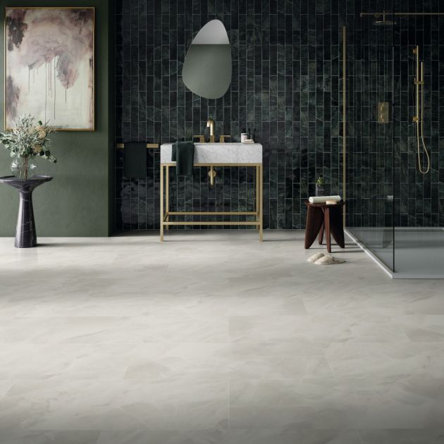 Bathroom with dark green wall with painting and side table and greenish-black tiles behind organic-shaped mirror, white and gold sink, and walk-in shower, with whitish-gray stone-look floors