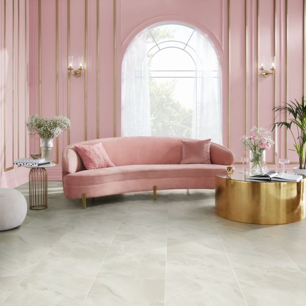 Living area with pink wainscoting, arched window, gold sconces, pink velvet sofa, and large gold coffee table topped with rose bouquet and wine glasses, with opalescent stone-look floor