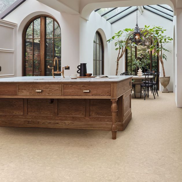 French country kitchen area, with exposed beams, arched doorframe, and large wooden island, with indoor trees flanking dining area in background, ecru wainscoting and taupe terrazzo floors