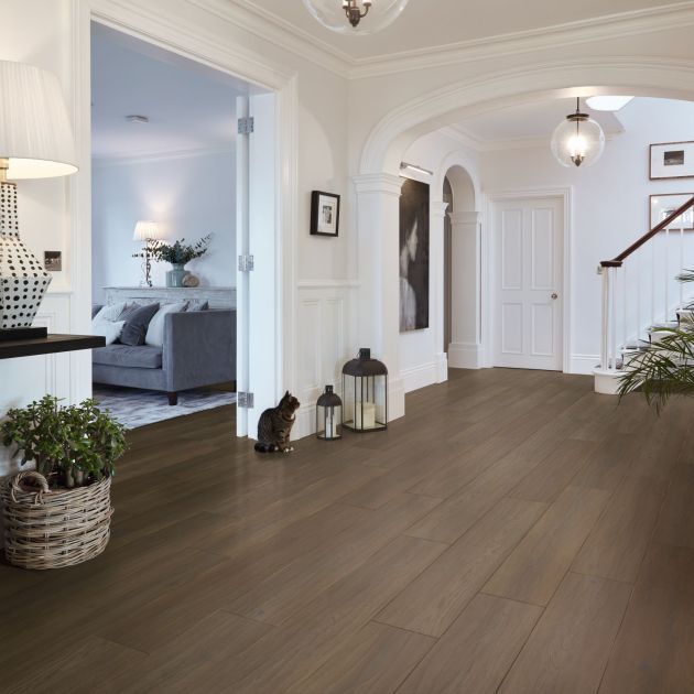 Refined and grounded hallway showing peek of seating area and staircase in background, with side table topped with dotted lamp, with potted plants, cat, and decorative candle lamps on chocolate-brown woodgrain floor