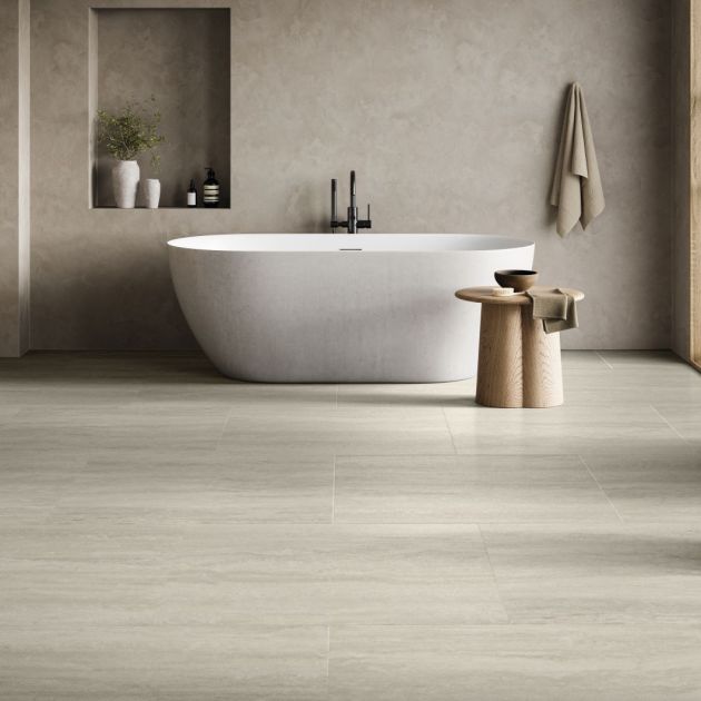 Bathroom featuring gray concrete-look wall with niche, hanging towel, and side table beside soaker bathtub, in room with gray travertine-style tile floor