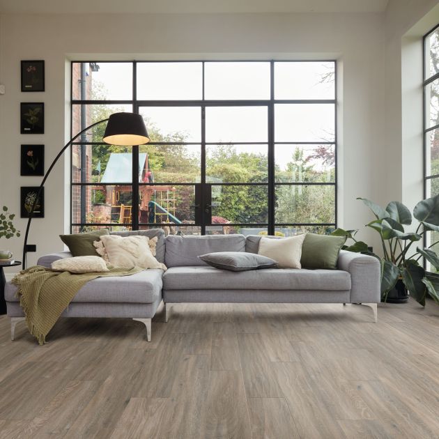 Living space with grayish-brown woodgrain floors, gray couch, arched floor lamp, and potted plant set before large windows with playground outside